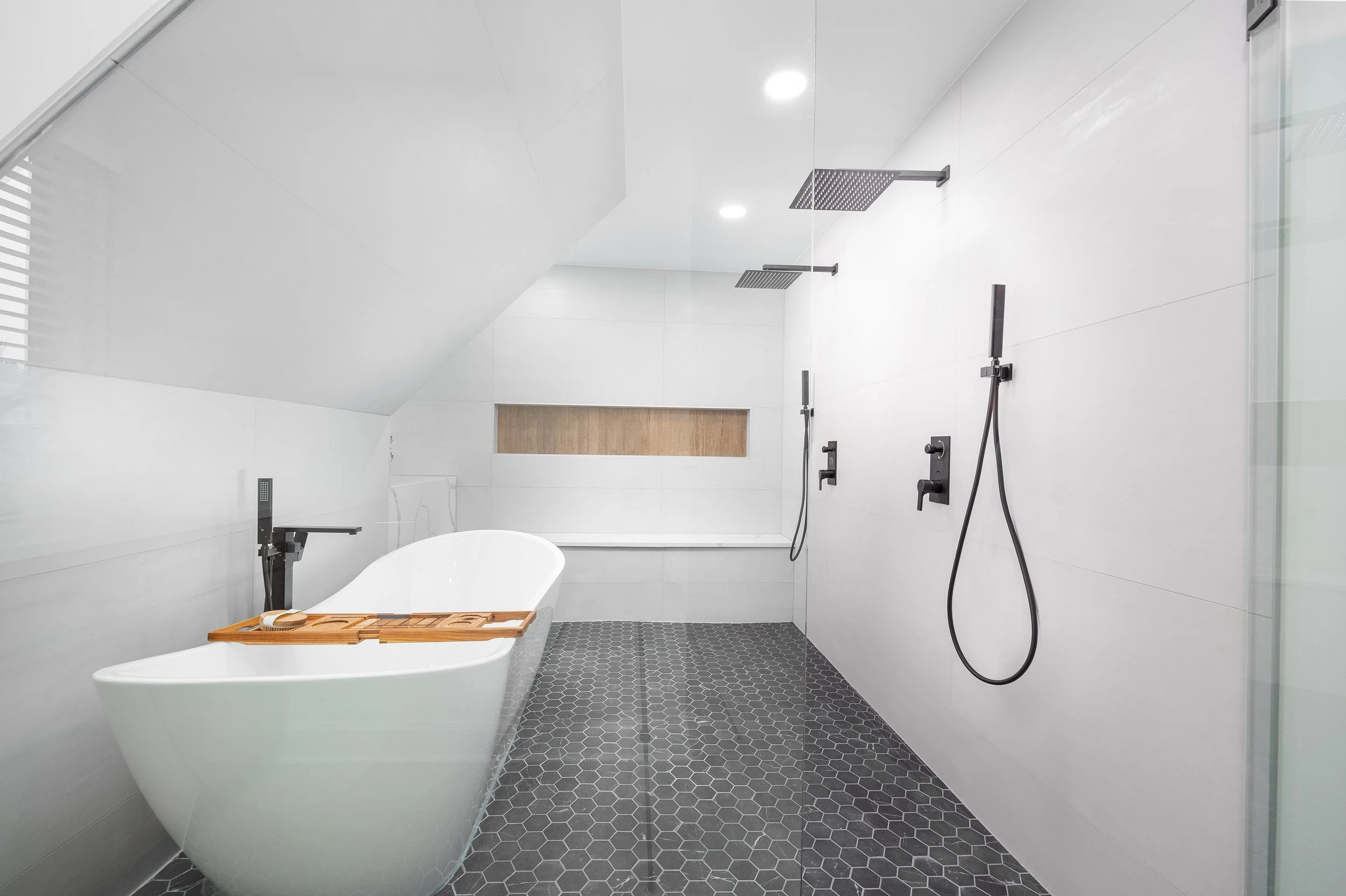 Wet room bathroom with large-format white tile, freestanding tub, twin rainfall showers and black hexagon floor tile